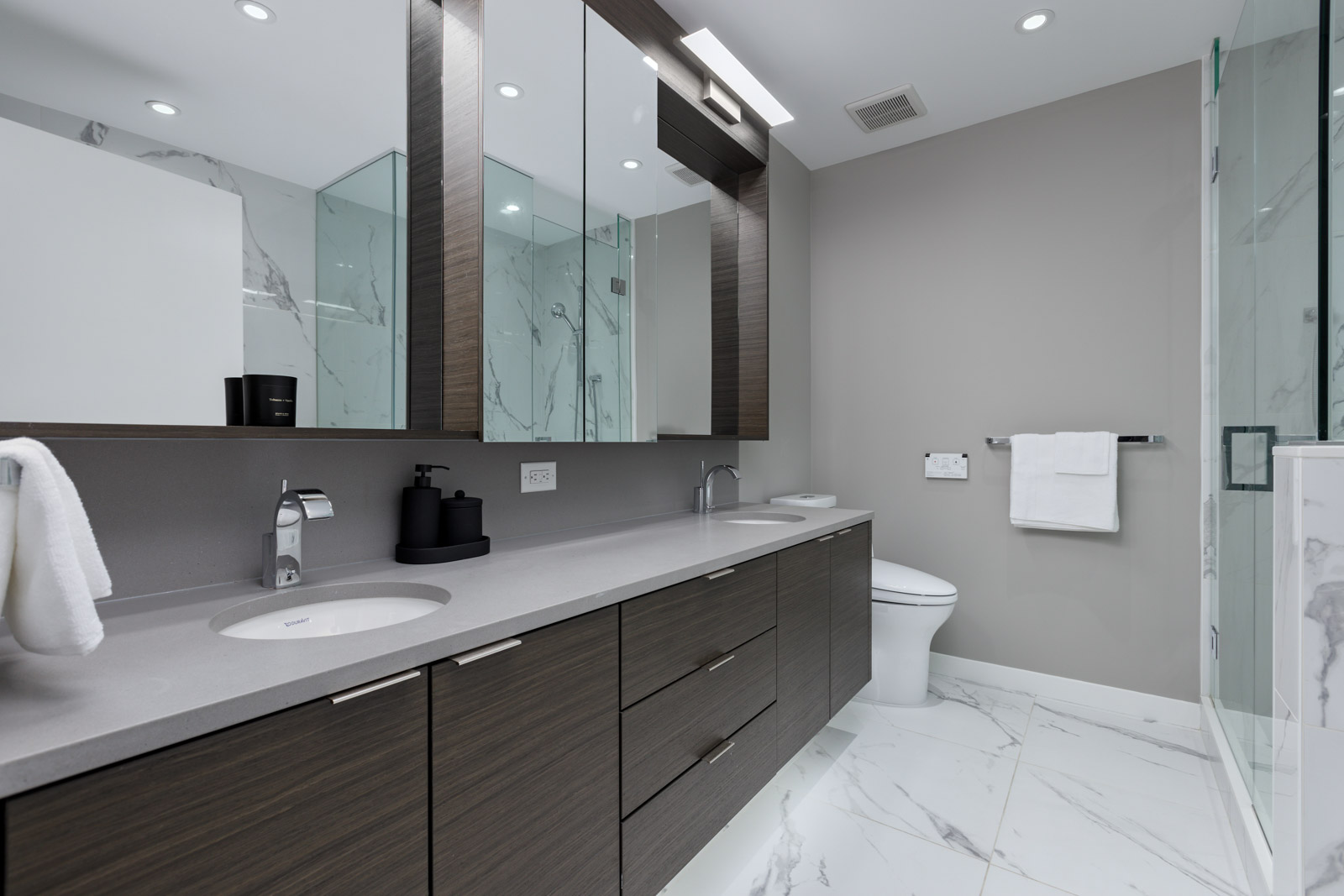 Modern bathroom with double sinks, dark wood cabinets, large mirrors, glass shower, wall-mounted toilet, towel rack, and white marble floor and walls.
