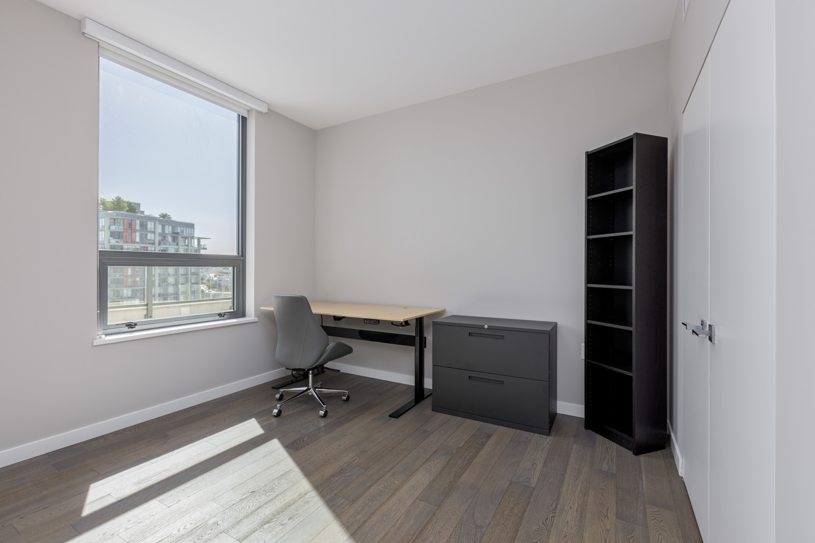 A modern, minimal office with a desk, chair, black file cabinet, and black bookshelf next to a large window with natural light and a city view.