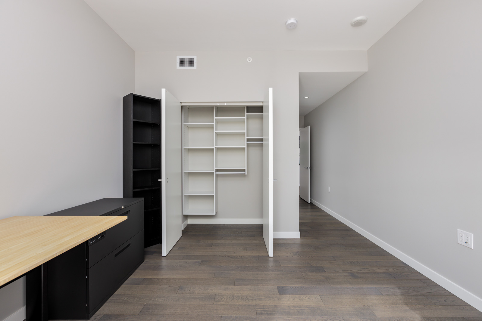 A modern, empty room with hardwood floors, open closet with shelves, a black desk, black shelving unit, and white walls.