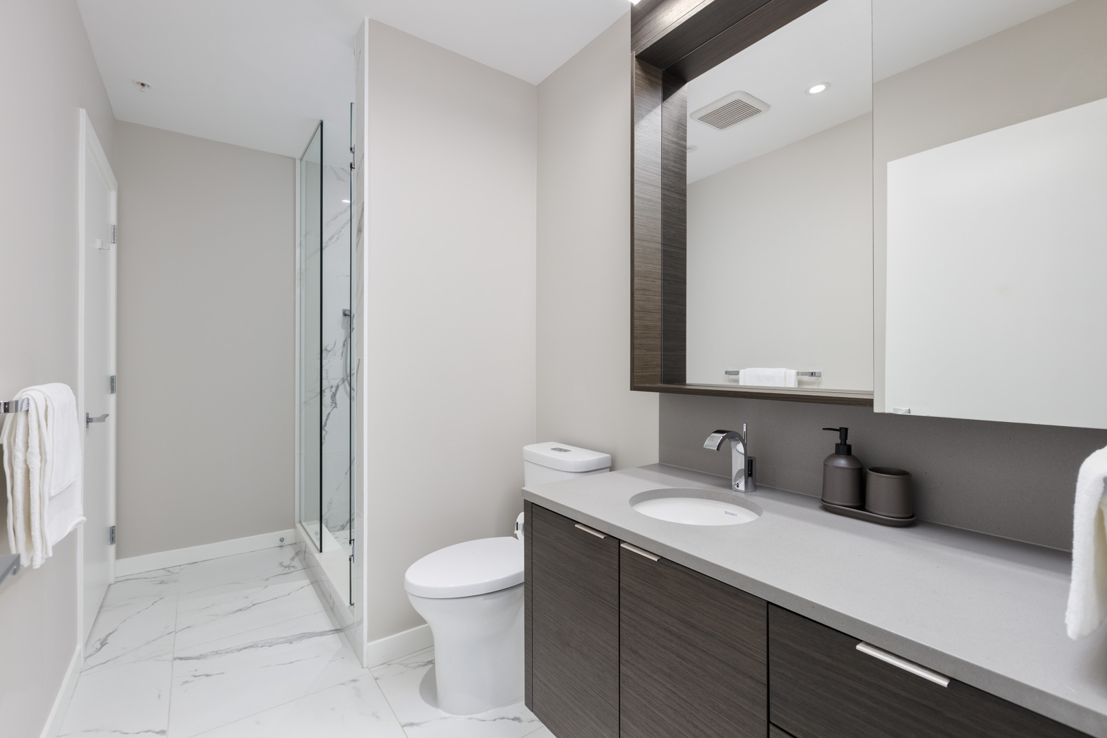 Modern bathroom with a glass shower, white toilet, dark wood vanity, large mirror, and marble tile floor.