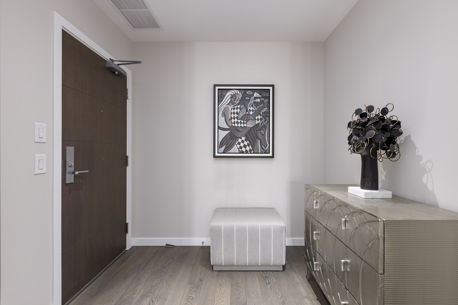A modern entryway with a closed wooden door, a patterned cabinet, a decorative black vase, a striped ottoman, and abstract artwork on a white wall.