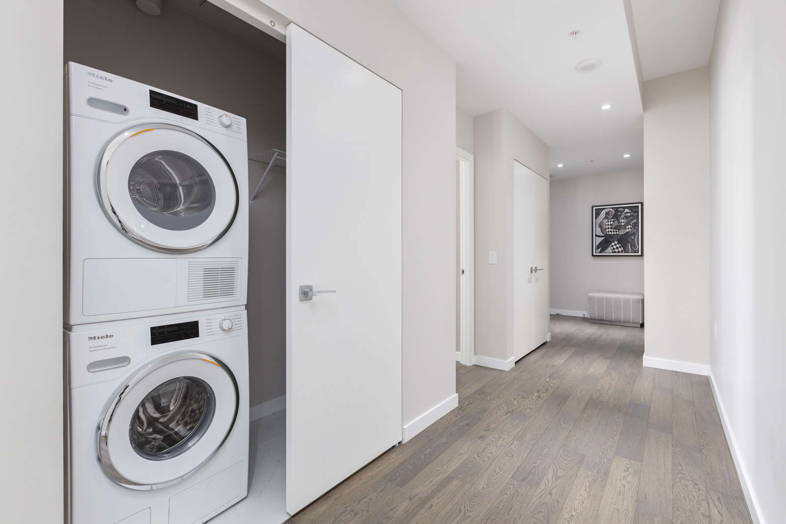 A hallway with a closet containing a stacked washer and dryer, partially open door, light-colored walls, wood floor, and artwork at the end of the hall.