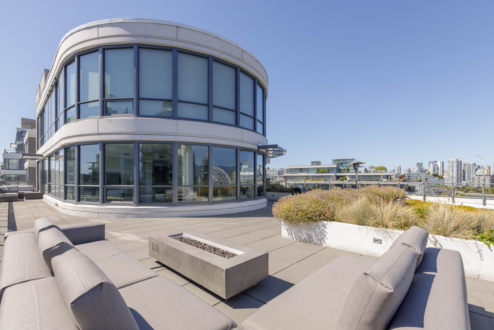 Modern rooftop terrace with outdoor seating, fire pit, and large curved glass windows; city buildings visible in the distance under a clear blue sky.