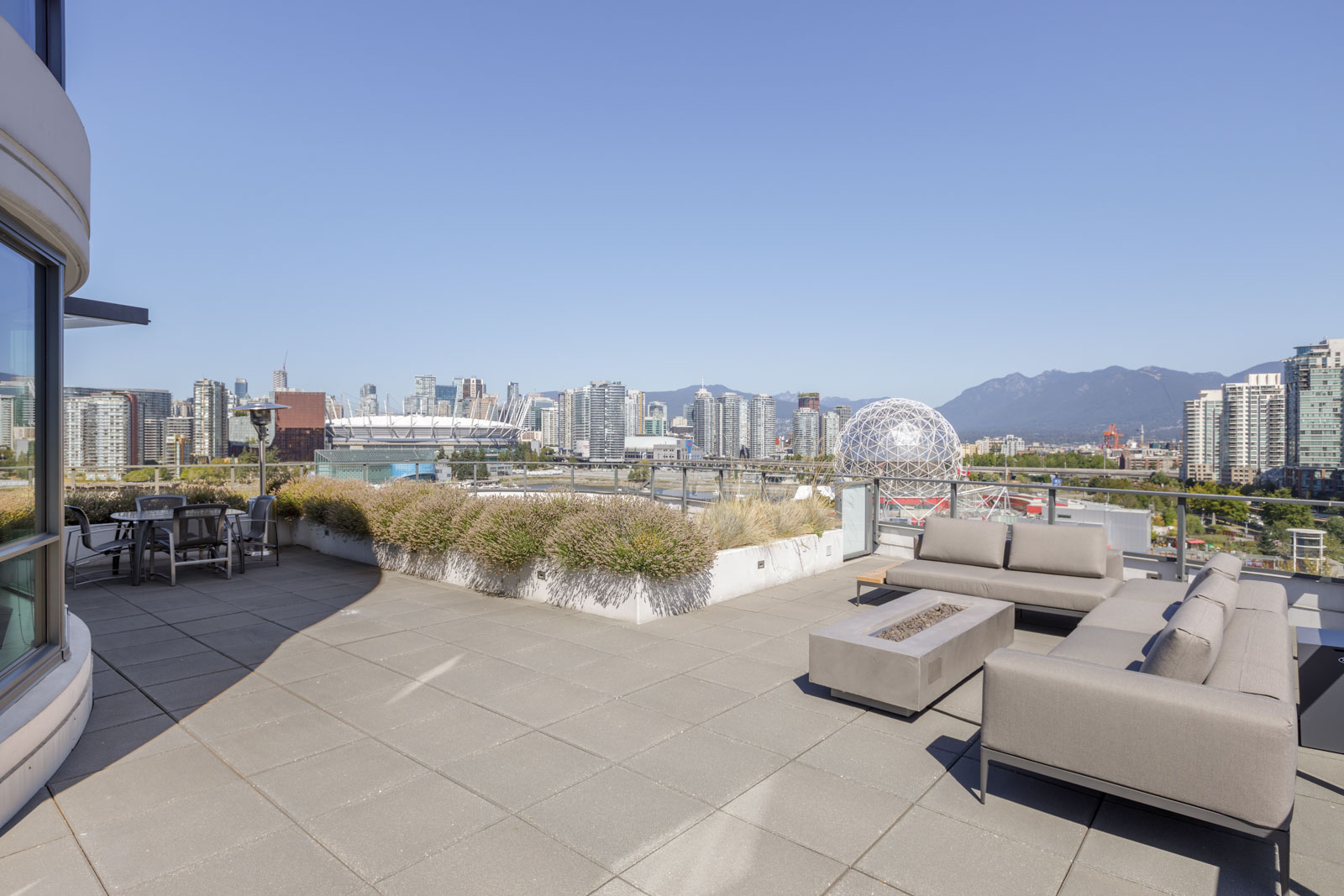 Spacious rooftop patio with modern seating, planters, and city skyline view featuring stadium, spherical structure, and mountains in the distance under a clear blue sky.
