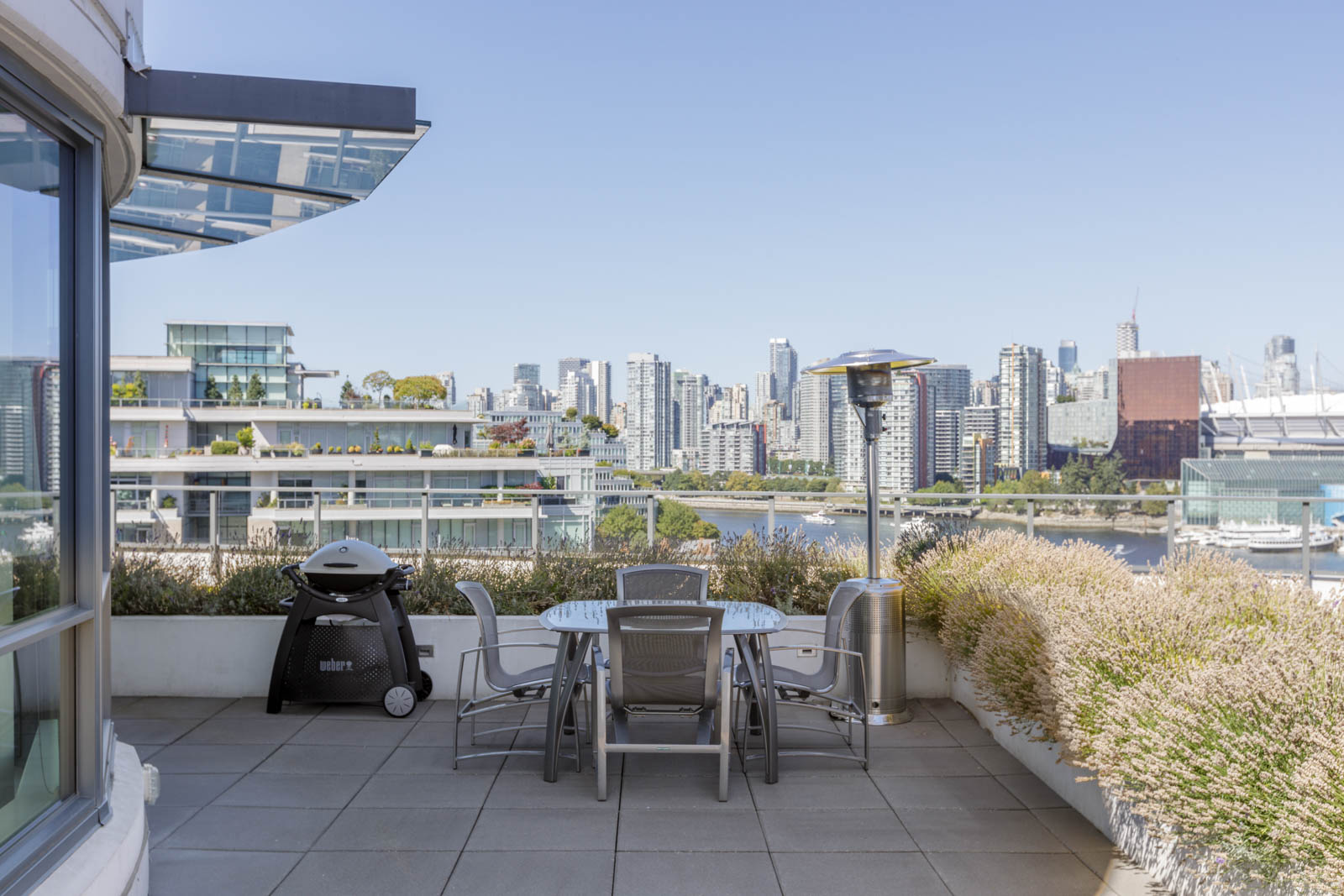 Outdoor patio with a table, chairs, grill, and heater overlooking a city skyline and waterfront on a clear day.