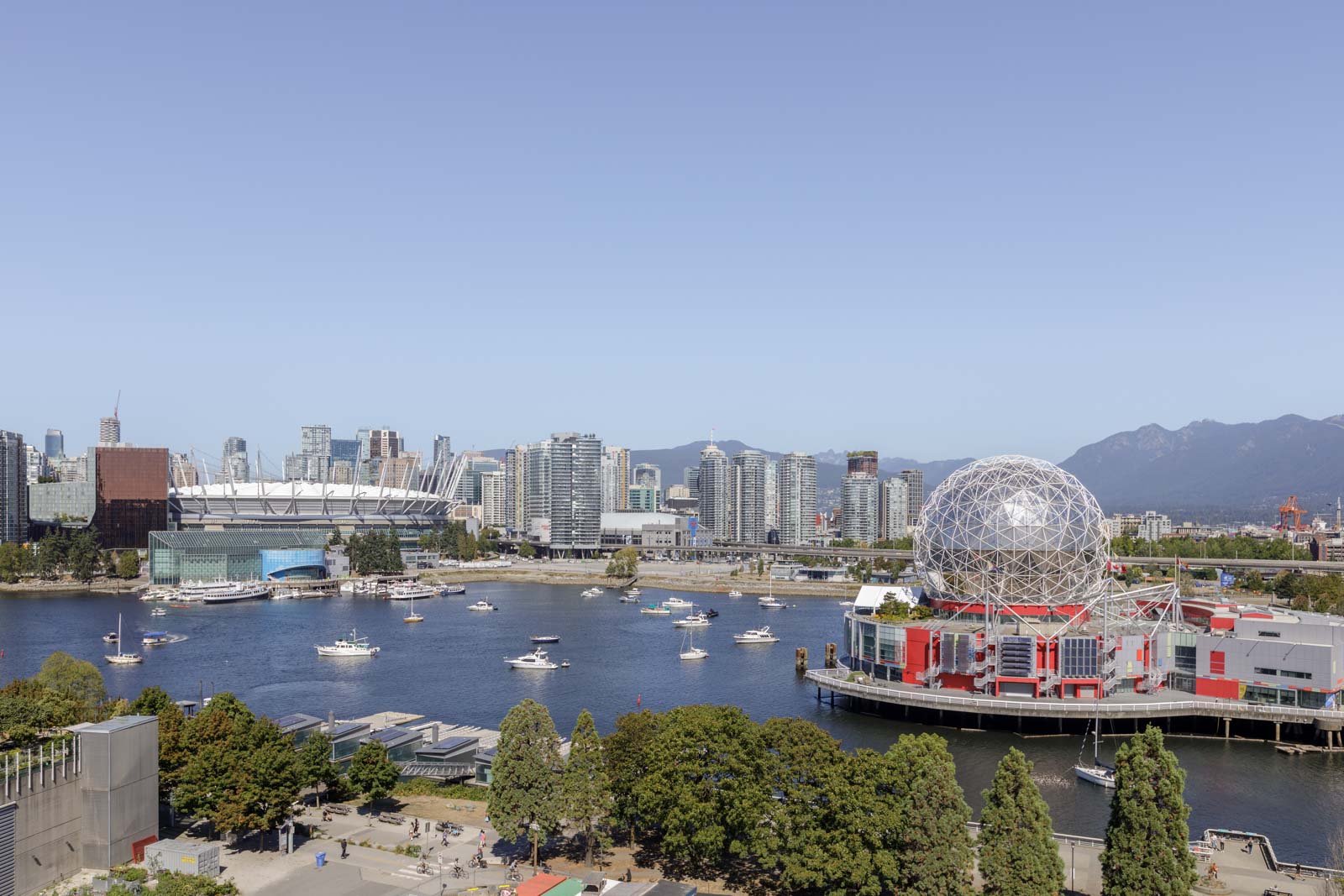 A waterfront cityscape of Vancouver, featuring Science World’s geodesic dome, BC Place Stadium, boats on the water, and glass high-rise buildings with mountains in the background.