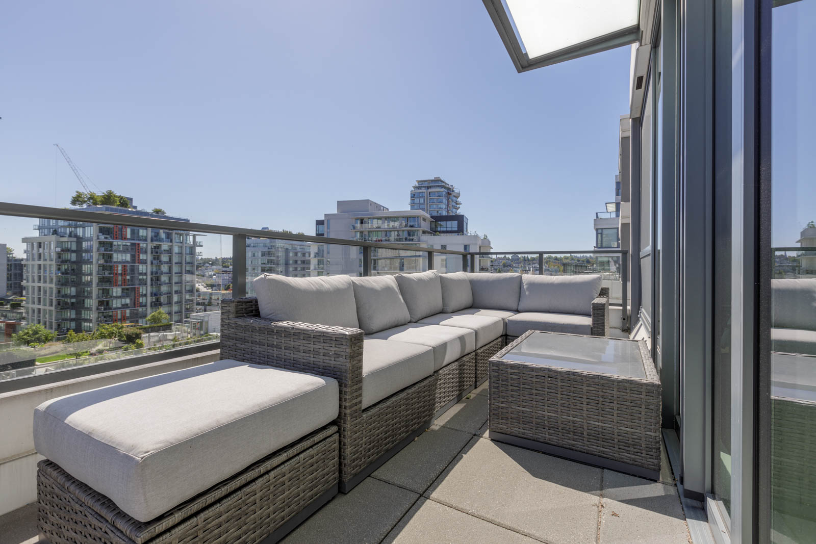Outdoor terrace with a gray wicker sectional sofa and glass-top table, surrounded by glass railing, overlooking modern city buildings under a clear sky.