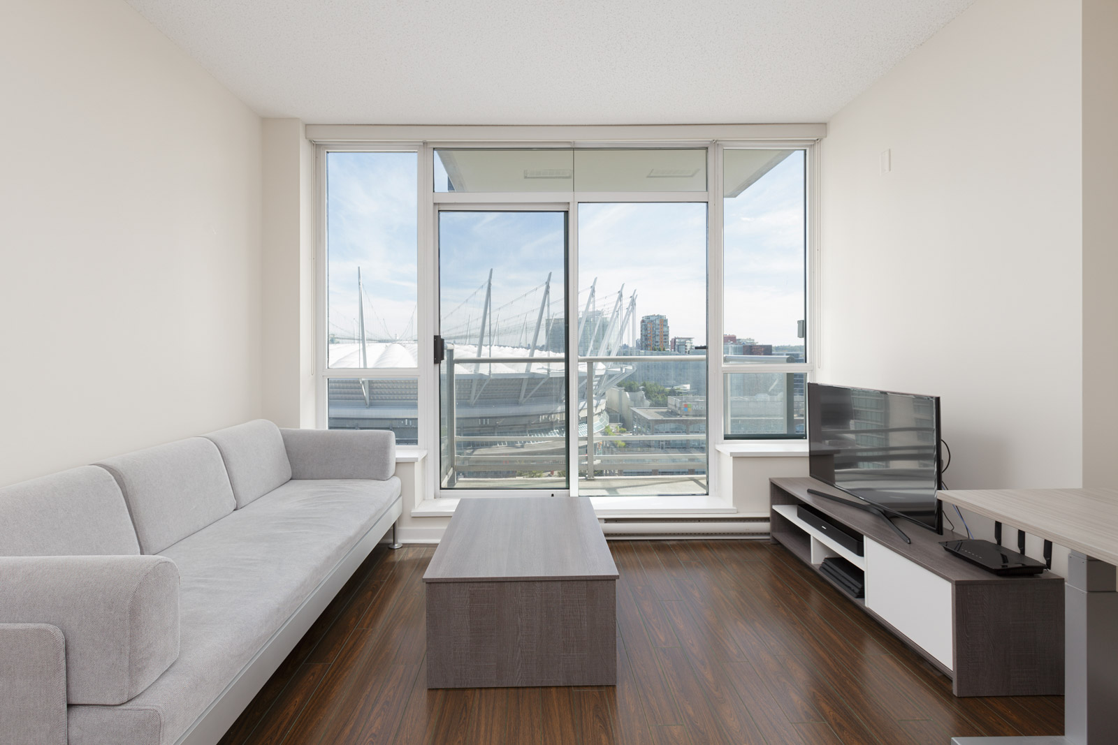 A modern living room with a gray sofa, wooden coffee table, TV on a stand, and large windows overlooking a cityscape and stadium.