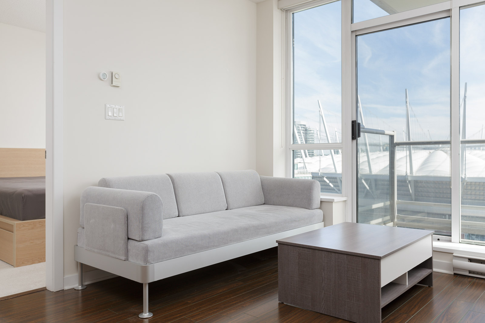 A modern living room with a gray sofa, wooden coffee table, large windows, and a view of a stadium roof outside. A bedroom is visible through an open doorway.