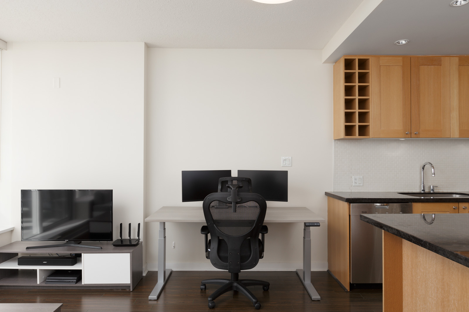 Modern home office setup with a desk, office chair, dual monitors, and a TV on a stand, adjacent to a kitchen area with wooden cabinets and a sink.