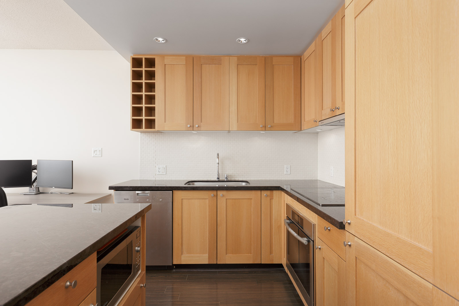 Modern kitchen with light wood cabinets, stainless steel appliances, a small wine rack, and a black countertop; a computer workstation is visible in the background.