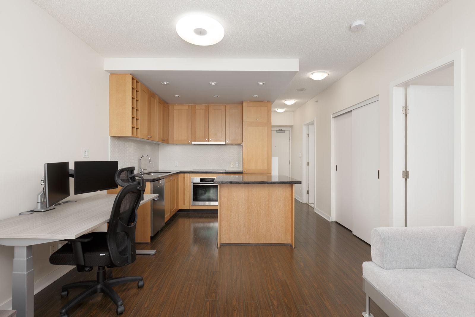 A modern studio apartment with a compact kitchen, wood cabinets, a work desk with dual monitors, a chair, and a light gray sofa on dark wood floors.