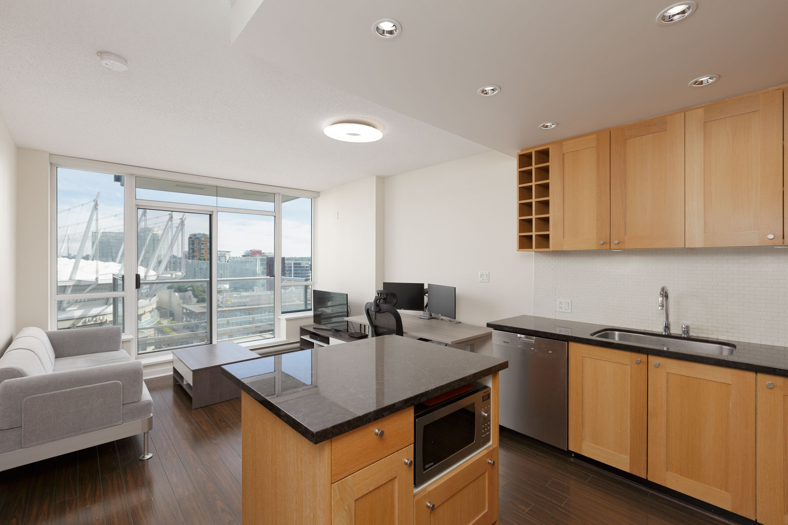 Modern apartment with light wood kitchen, island, and open living area featuring a sofa, desk with dual monitors, large window, and city view in the background.