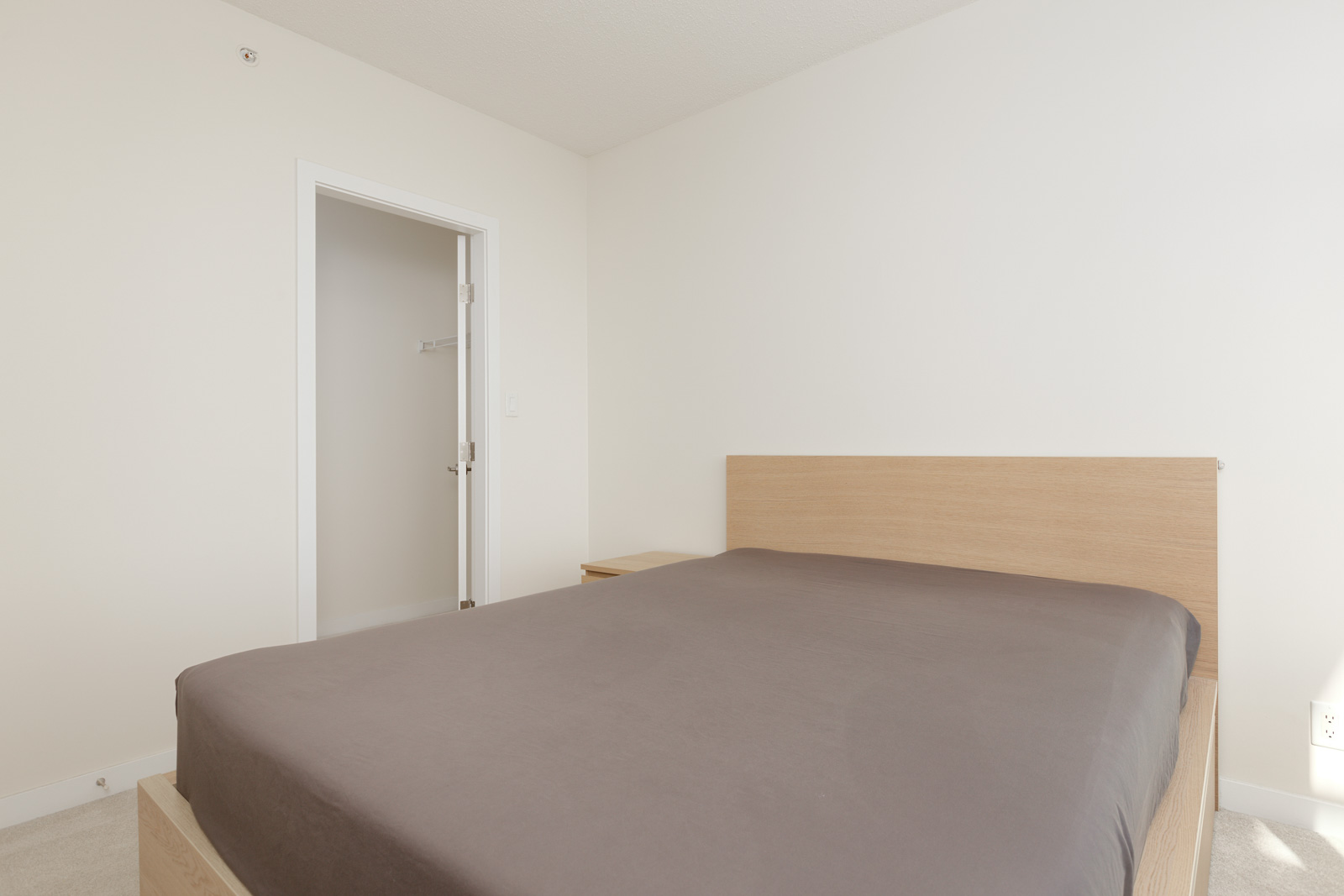 A minimalist bedroom with a wooden bed frame, gray bedspread, small nightstand, and an open door leading to a closet.