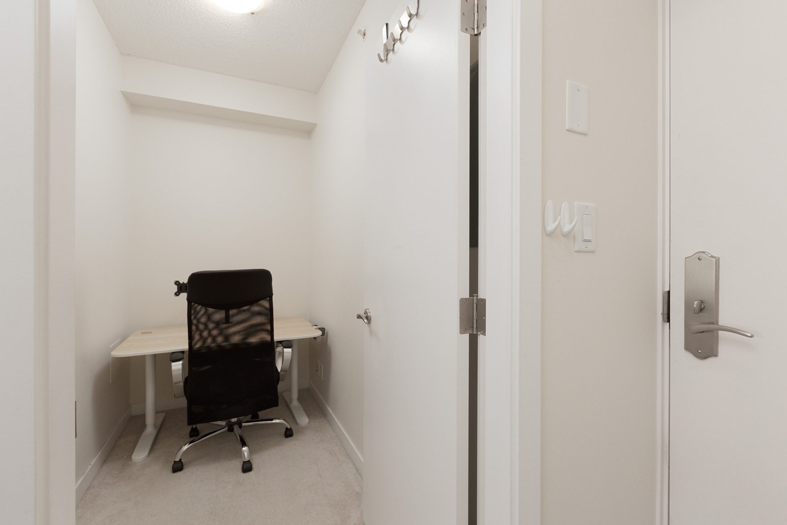 A small, windowless room with a white door, a black office chair, and a light-colored desk against the far wall under a ceiling light.