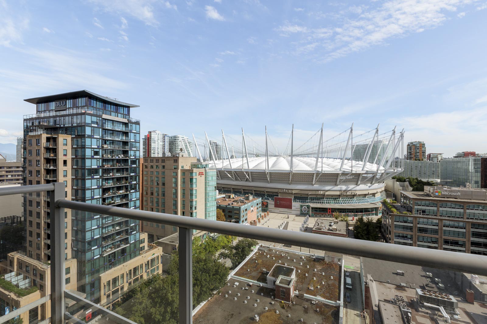 View from a balcony overlooking modern buildings and a large stadium with white roof and spires under a partly cloudy sky.