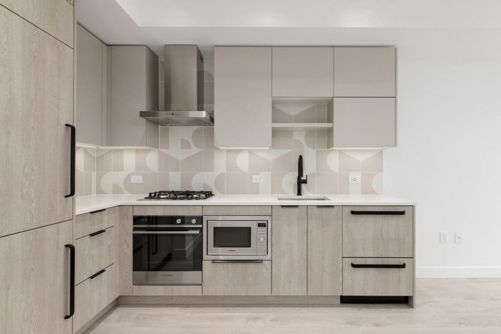 Modern kitchen with light wood cabinets, geometric backsplash, built-in oven, microwave, gas stove, and black fixtures under bright lighting.
