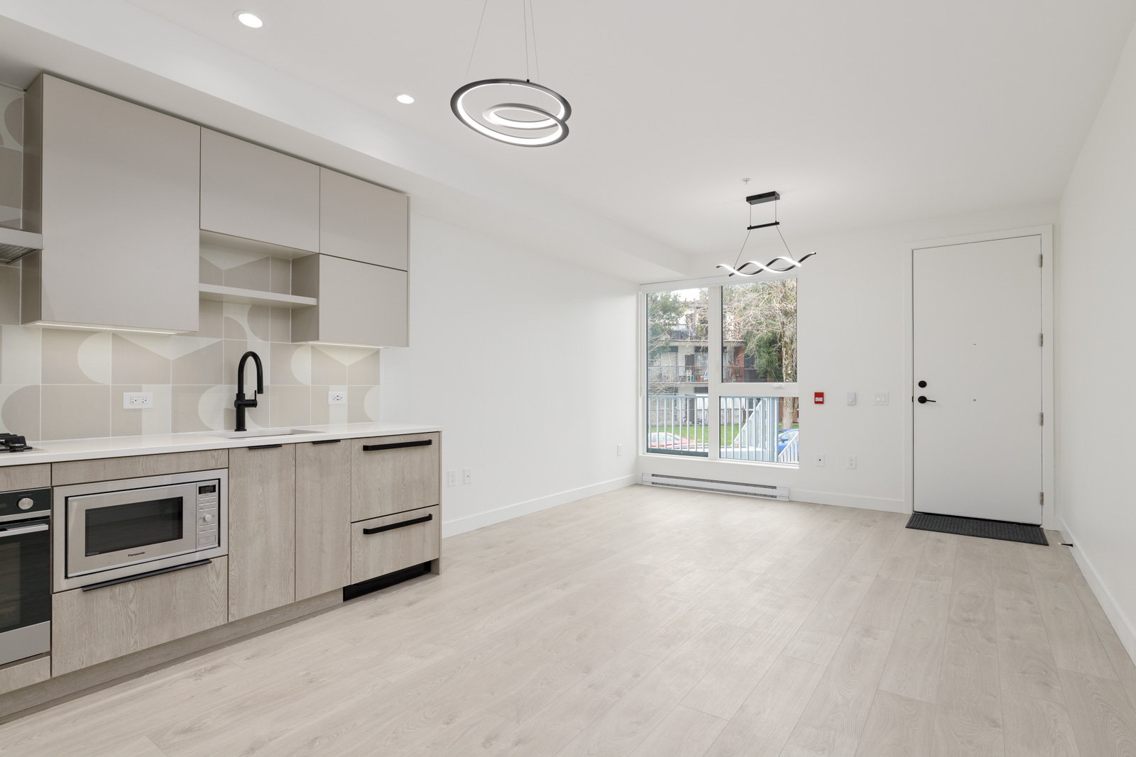 Modern, unfurnished apartment kitchen and living area with light wood floors, built-in appliances, white walls, large window, and contemporary lighting fixtures.