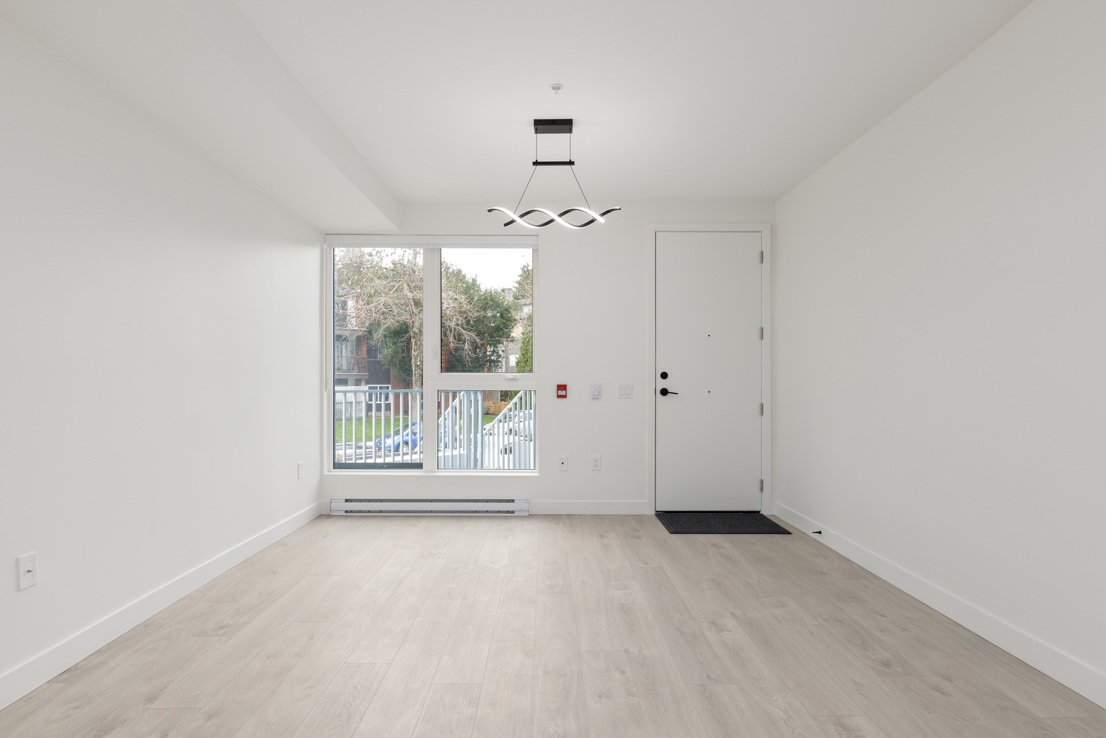 Empty, modern room with light wood flooring, large window, white walls, a black front door mat, and a contemporary ceiling light fixture.