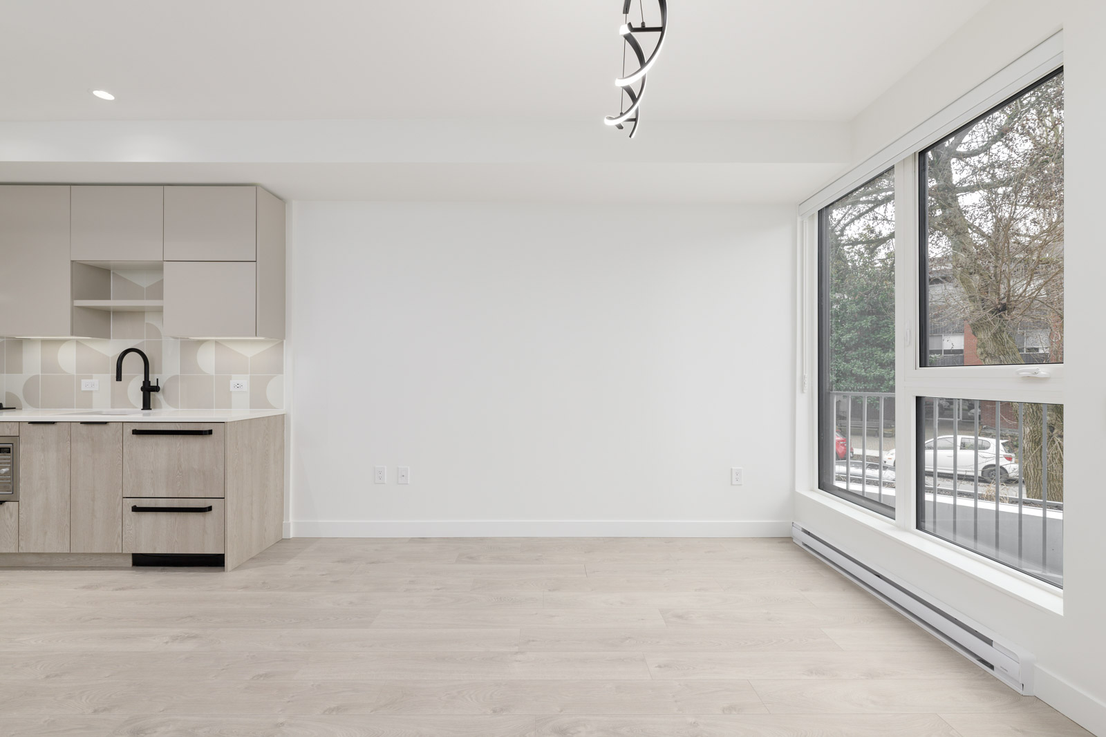 Bright, modern unfurnished room with light wood flooring, large window, and a minimalist kitchen area featuring neutral cabinets and geometric backsplash tiles.