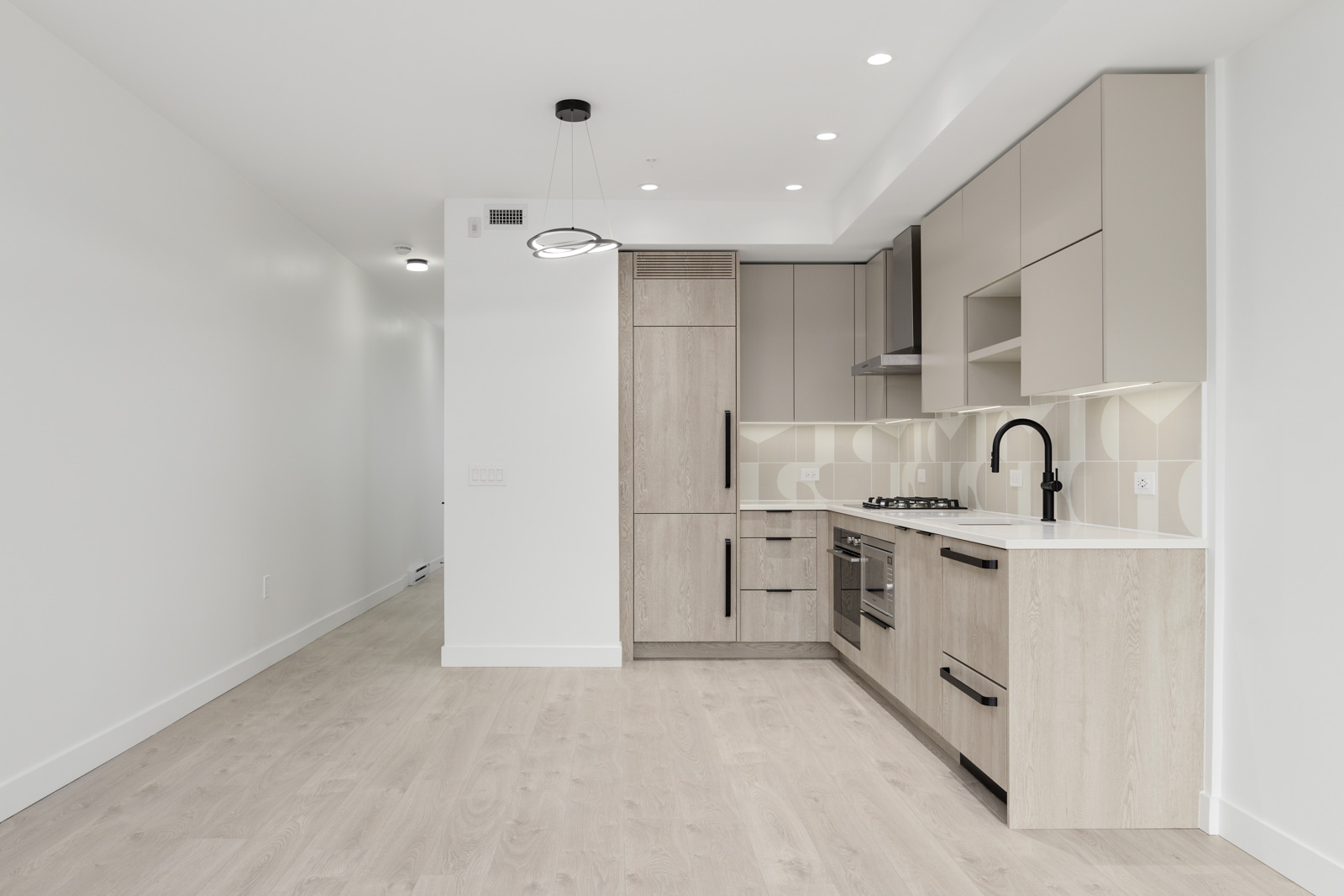 Modern kitchen with light wood cabinetry, built-in appliances, white countertops, and geometric backsplash, adjacent to an empty open living area with light wood flooring and white walls.