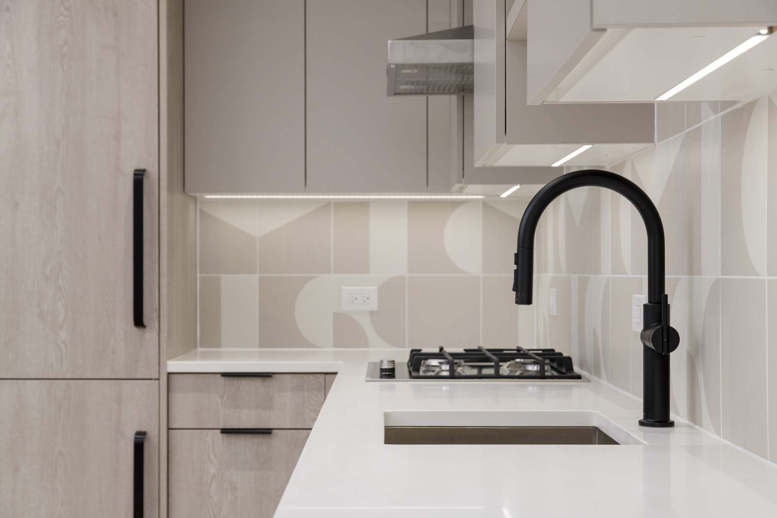 Modern kitchen with light wood cabinets, white countertops, black faucet, built-in gas stove, and geometric patterned tile backsplash.