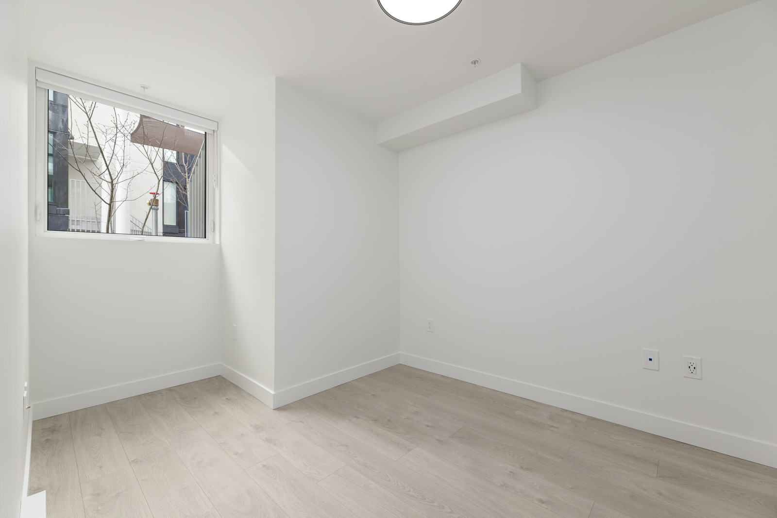 Empty, unfurnished room with light wood flooring, white walls, a single window on the left, and electrical outlets on the right wall. Natural light enters through the window.