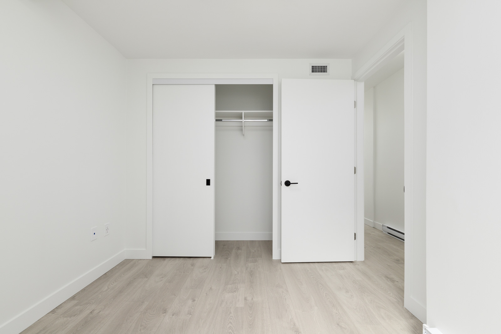 Empty room with light wood flooring, white walls, an open closet with sliding doors, and a white door leading to another space.