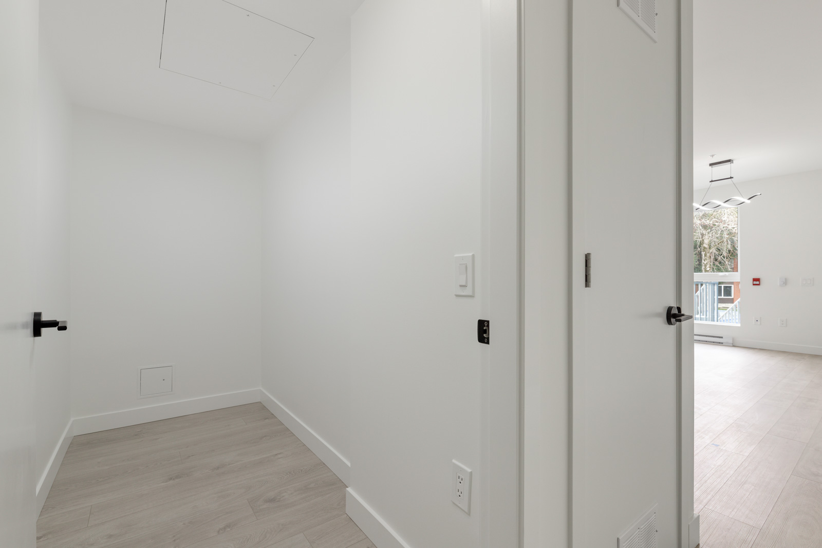 A small empty white room with light wood flooring, a closed vent on the ceiling, and an open door leading to a brighter living area.