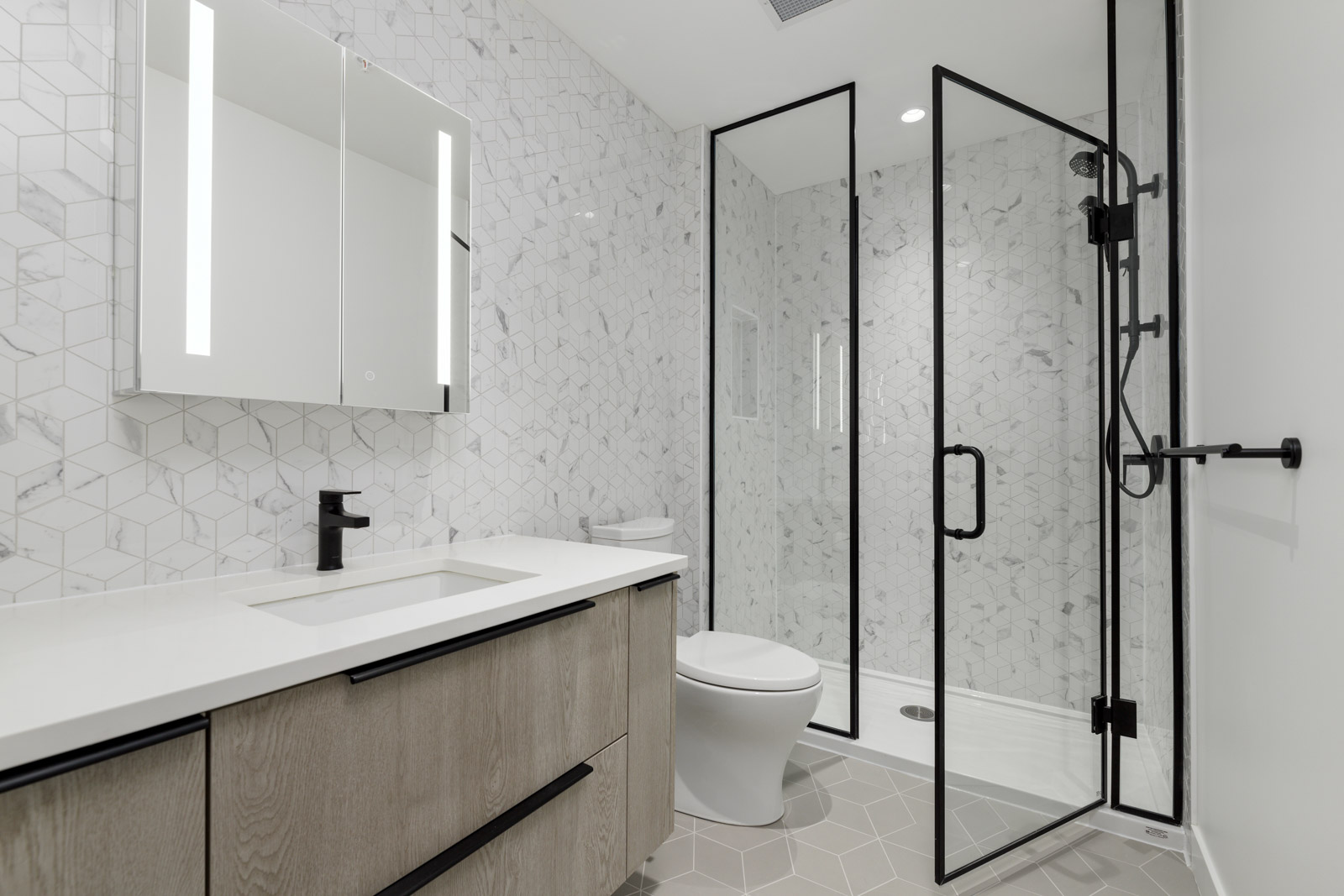 Modern bathroom with light wood vanity, white countertop, wall-mounted mirror, toilet, and glass-enclosed shower. Walls and floor feature geometric tile patterns.