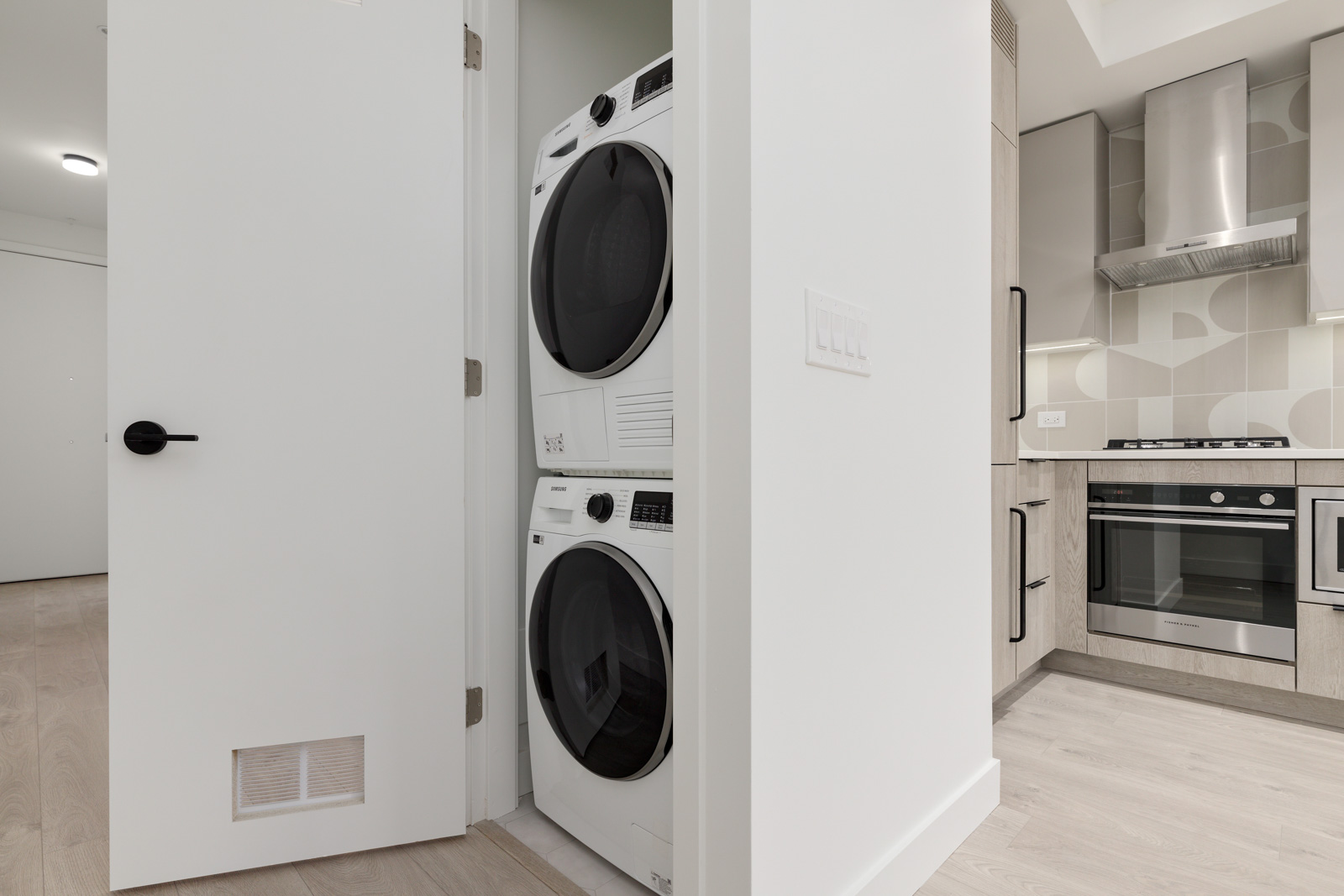 Stacked washer and dryer in a small laundry closet next to a modern kitchen with light wood floors and stainless steel appliances.