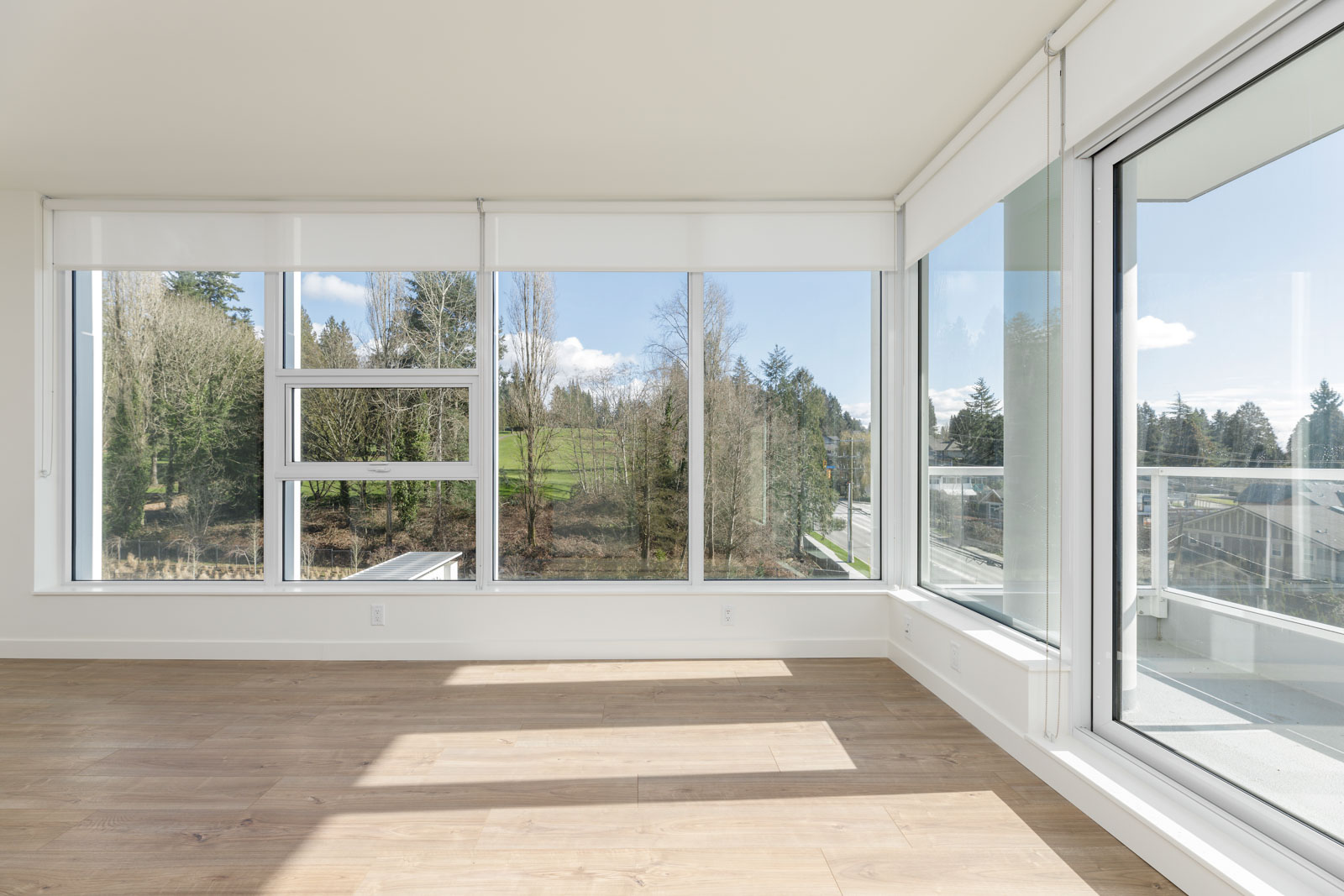 Bright, empty room with large floor-to-ceiling windows, white walls, and light wood flooring, overlooking trees and a grassy area outside.
