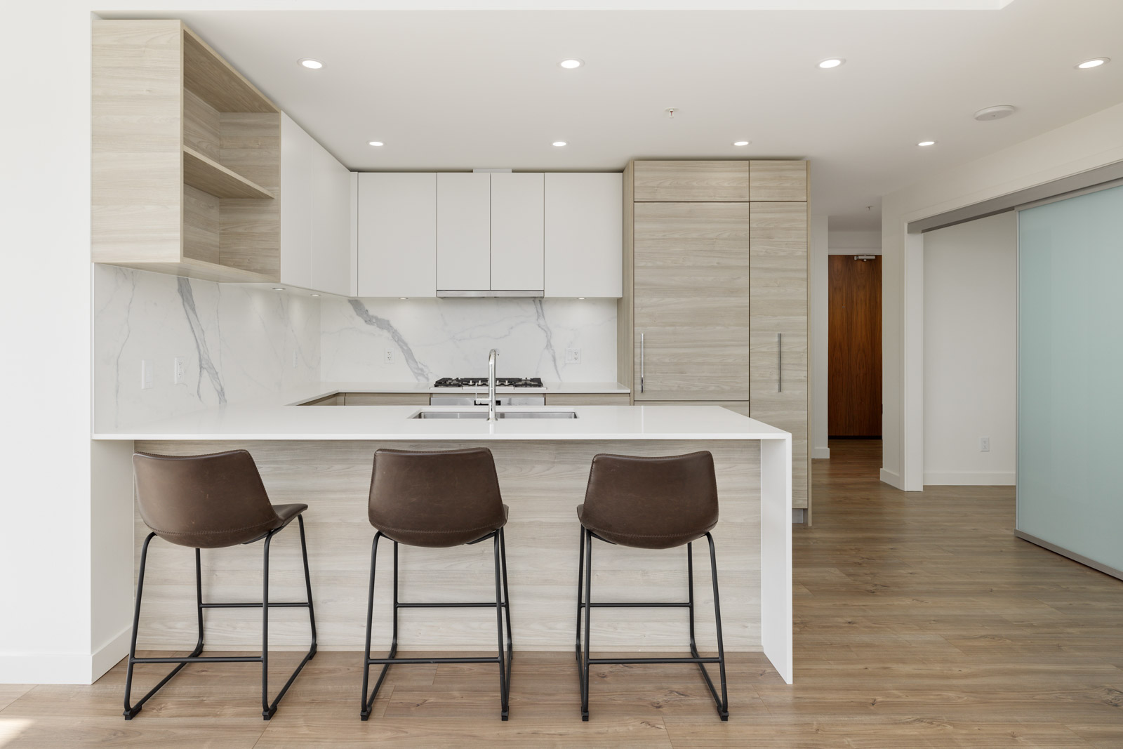 Modern kitchen with light wood cabinets, white countertops, marble backsplash, and three brown barstools at an island.