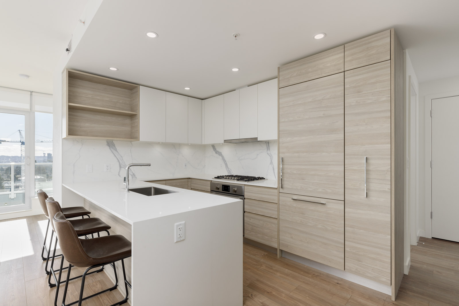 Modern kitchen with light wood cabinets, white countertops, built-in appliances, a central island with sink, and three brown barstools. Large windows let in natural light.