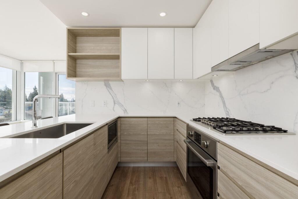 Modern kitchen with light wood cabinets, white countertops, built-in appliances, gas stove, and marble backsplash, featuring large windows with natural light.