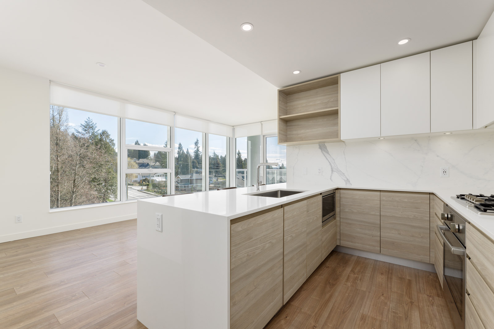Modern kitchen with light wood cabinetry, white countertops, built-in appliances, and large windows providing natural light.