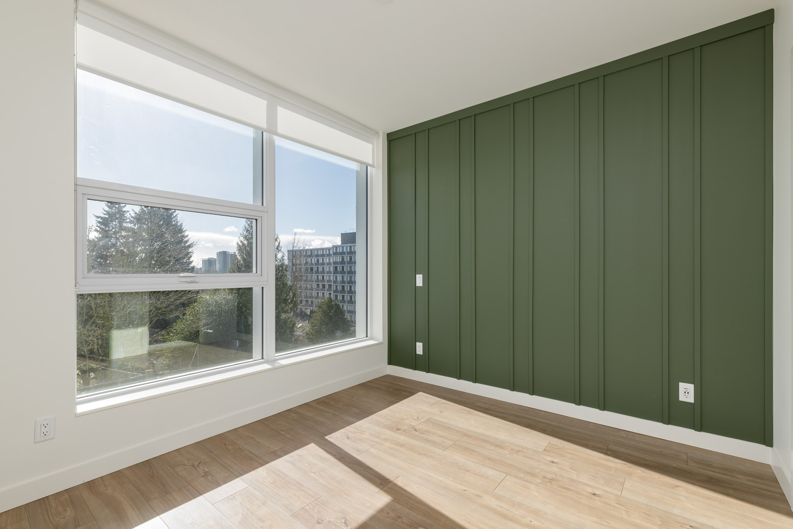 Empty room with large windows, a green accent wall, and light wood flooring. Sunlight enters, casting shadows on the floor. Trees and a building are visible outside.