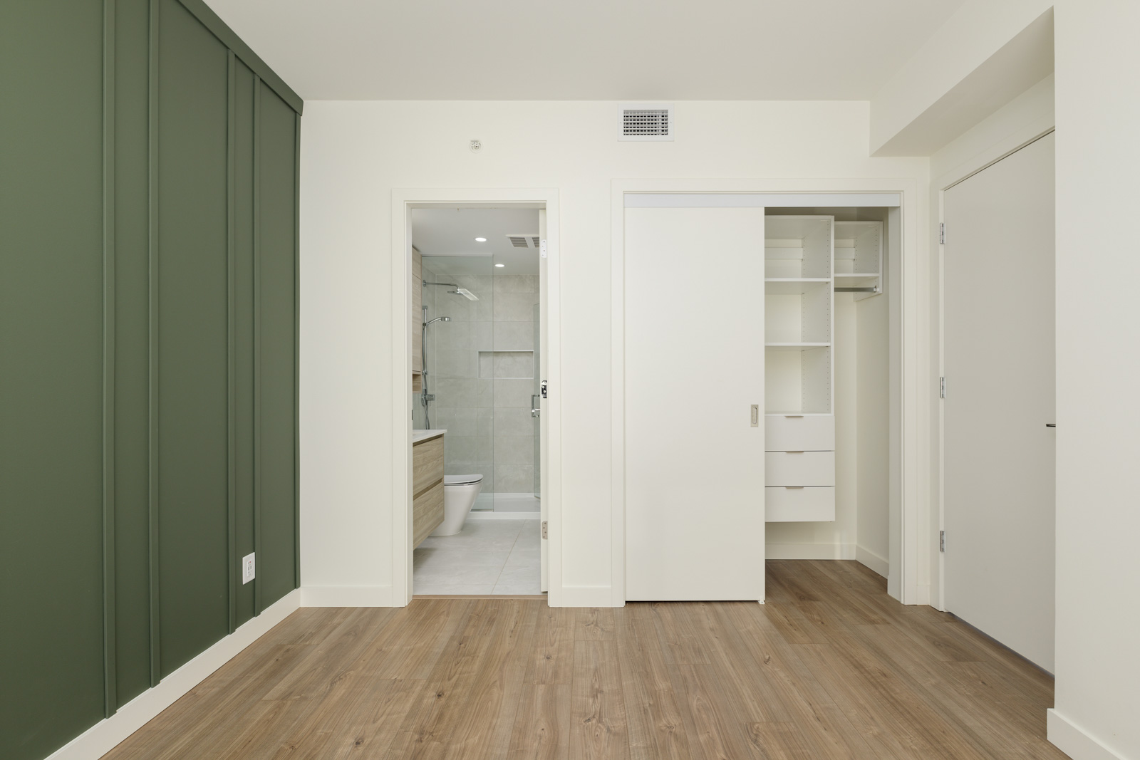 A modern, empty bedroom with wood flooring, a green accent wall, an open closet with shelves, and an en-suite bathroom visible through an open door.