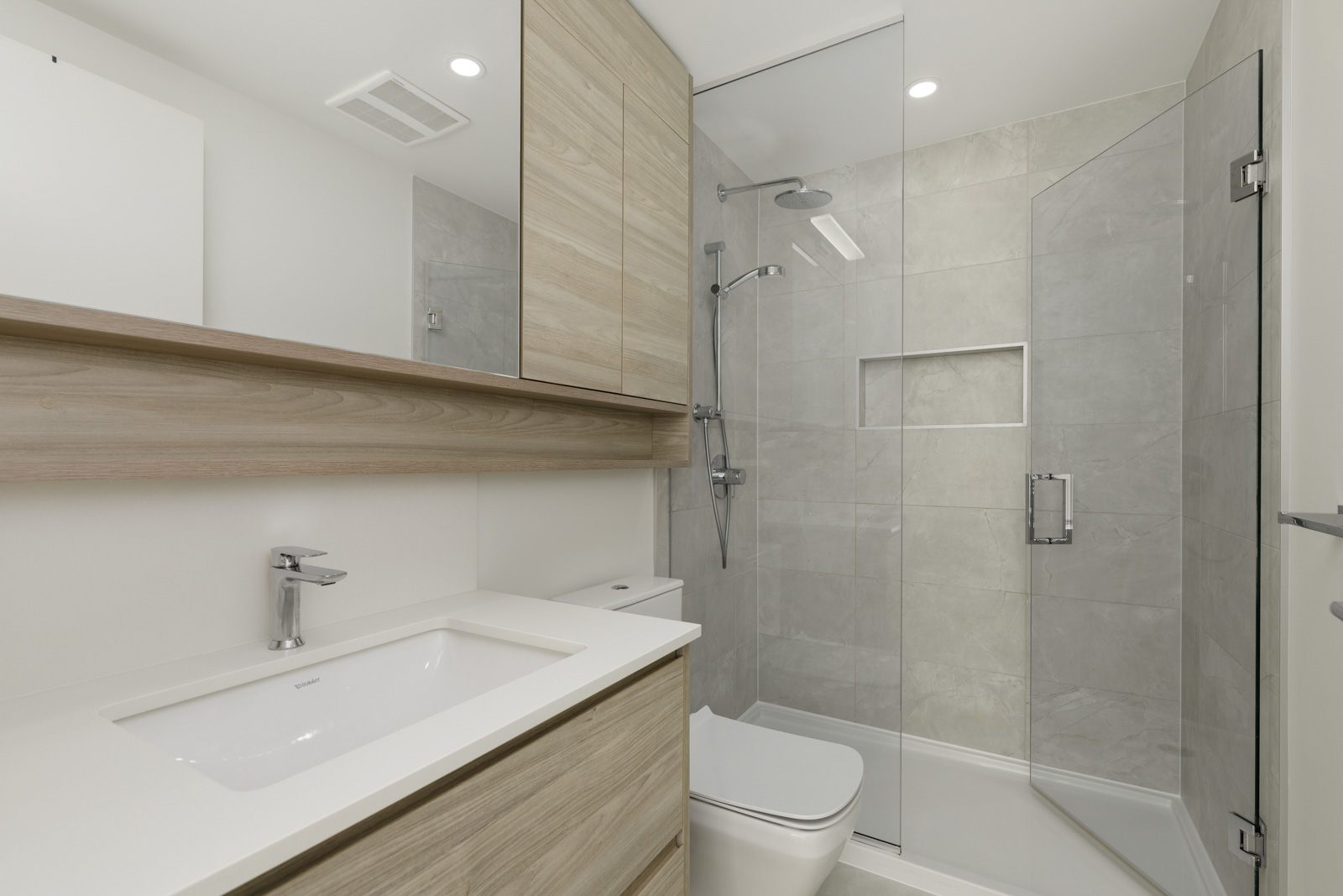 Modern bathroom with light wood cabinetry, a glass-enclosed shower, built-in shelves, wall-mounted sink, and neutral-colored tiles.