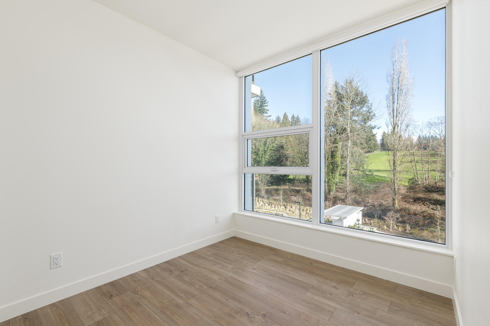 Empty room with white walls, wood flooring, and large floor-to-ceiling windows offering a view of trees and a grassy landscape outside.