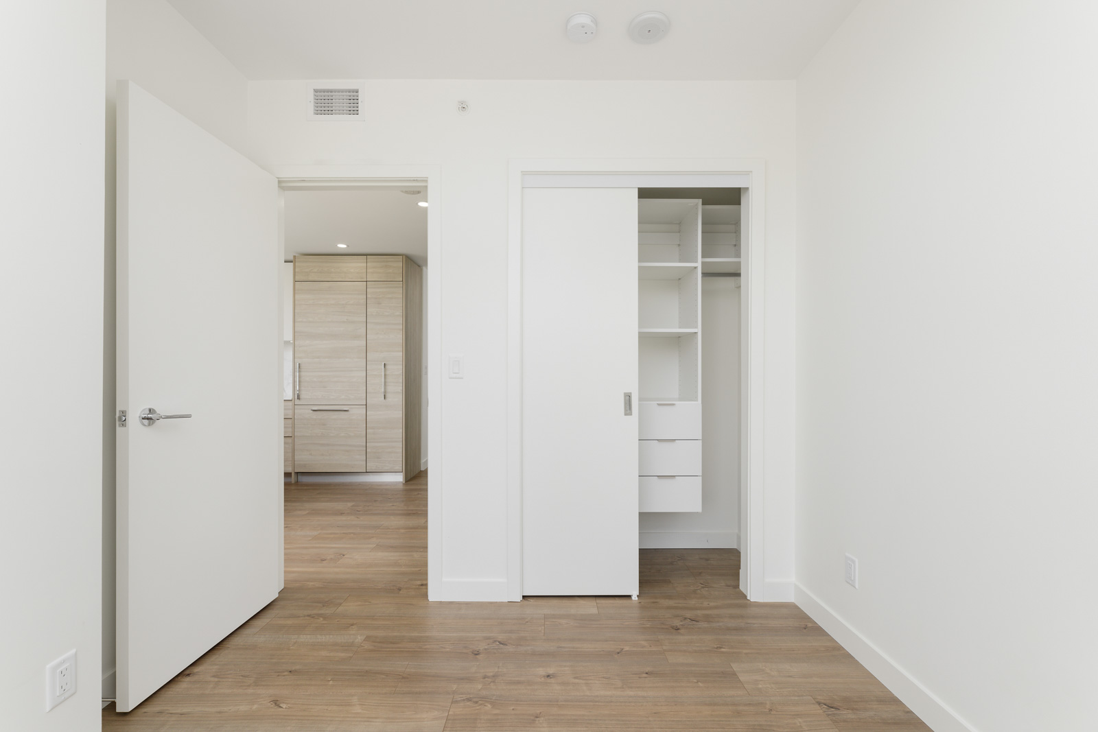 Empty room with wooden floors, white walls, an open door, and a closet with sliding doors and built-in shelves.