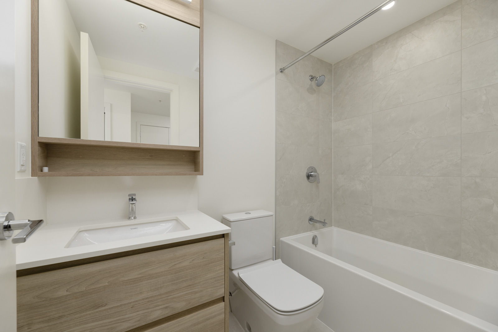 Modern bathroom with light wood vanity, white countertop, wall-mounted mirror cabinet, toilet, and bathtub with beige tiled shower walls.