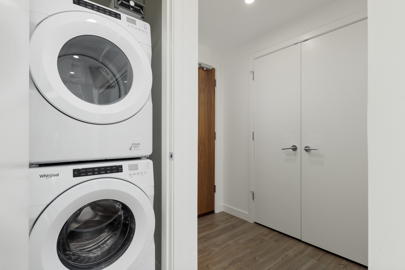 A stacked washer and dryer are installed in a closet next to double white doors in a modern, clean laundry area with wood flooring.