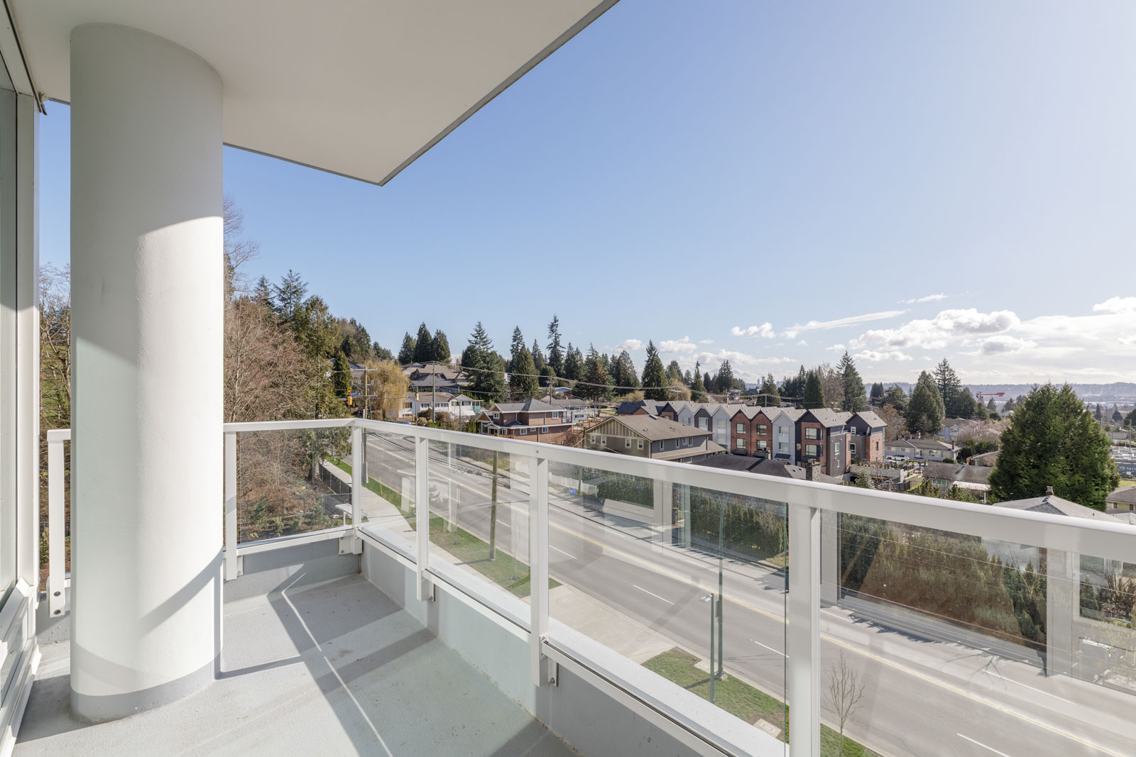 A modern apartment balcony with glass railing overlooks a suburban neighborhood, trees, and a clear blue sky on a sunny day.
