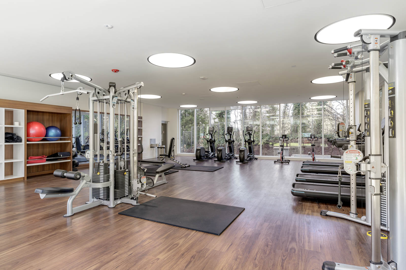 Modern gym with wooden floors, weight machines, treadmills, exercise bikes, and shelving with fitness balls; large windows provide outdoor view and natural light.