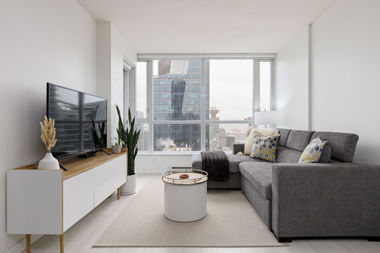 Modern living room with a grey sectional sofa, patterned cushions, TV on a wooden stand, potted plants, round coffee table, and large windows showing a cityscape view.