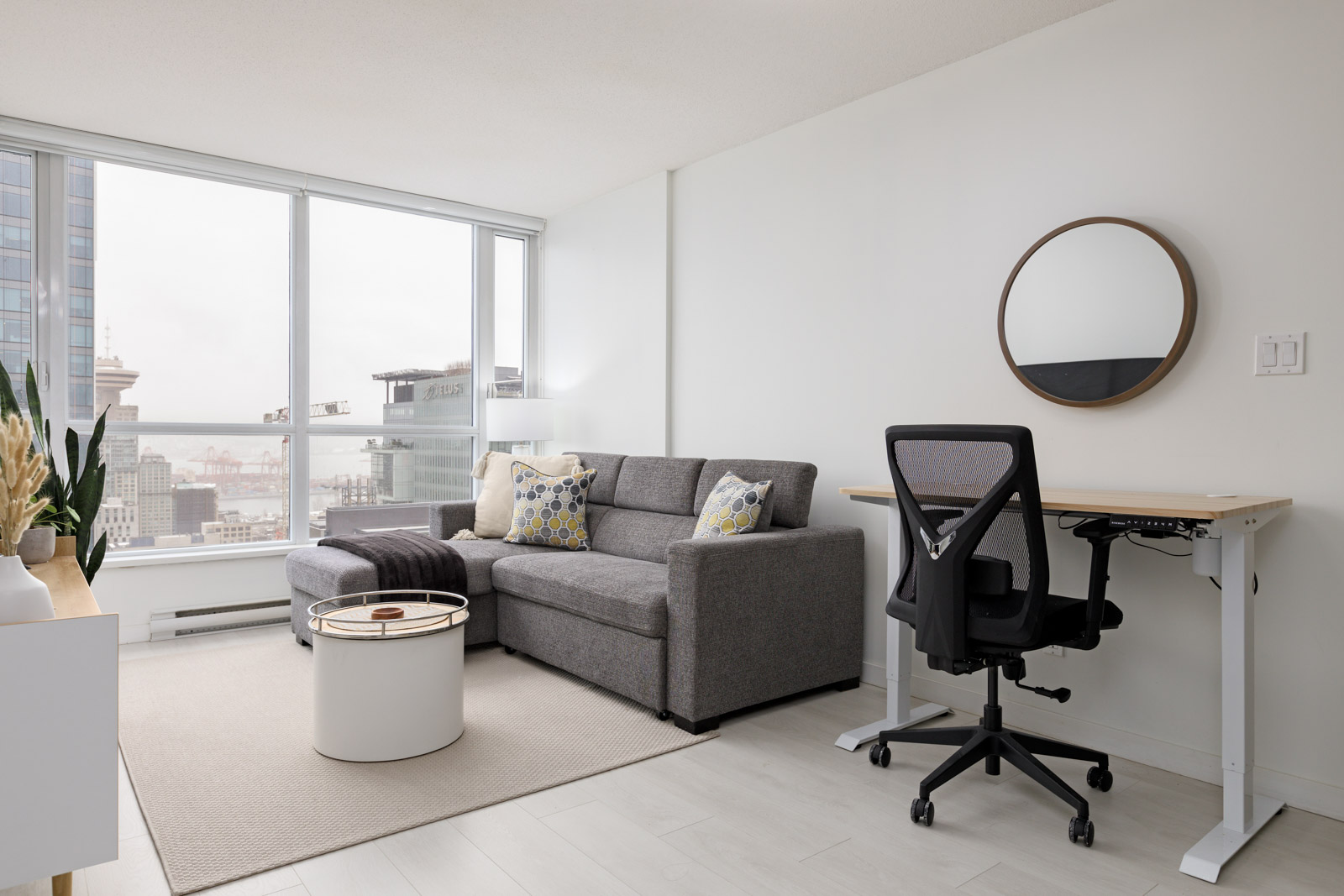 A modern apartment living area with a grey sectional sofa, round coffee table, workspace with office chair, and large windows overlooking a cityscape.