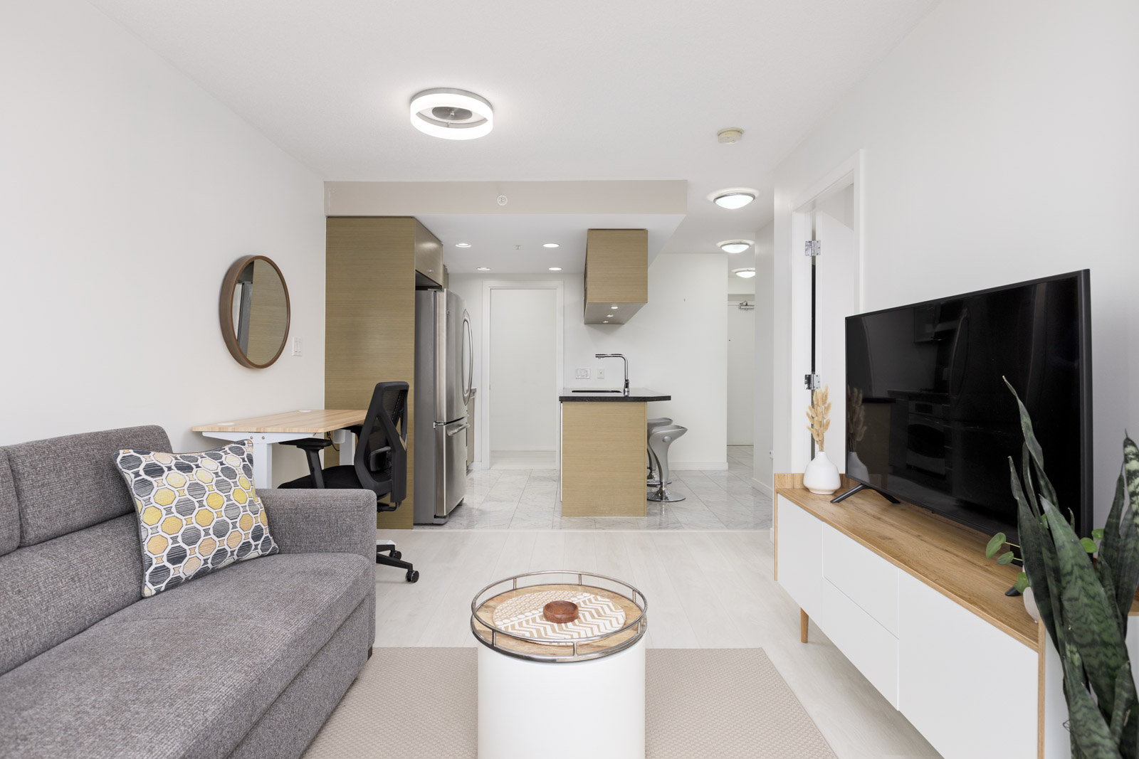Modern open-plan living room and kitchen with a gray sofa, round coffee table, TV on a wooden stand, office desk, and neutral-colored decor.
