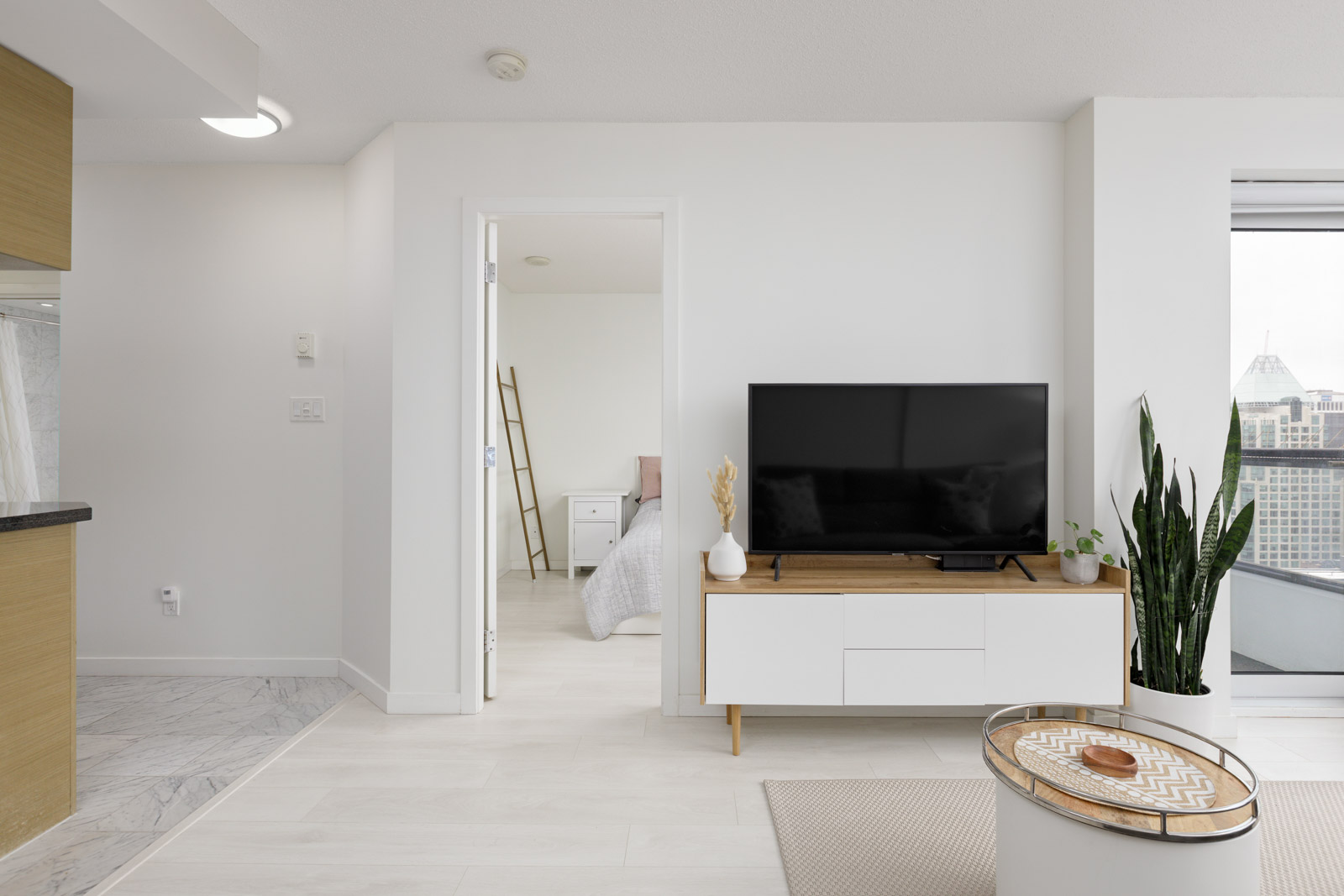 Modern living room with a flat-screen TV on a white console, a potted plant, and a view into a bedroom; cityscape visible through sliding glass door.