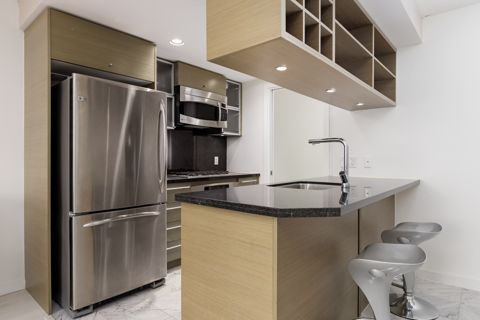 Modern kitchen with stainless steel appliances, light wood cabinets, black countertops, built-in shelving, and two gray bar stools at a peninsula with a sink.